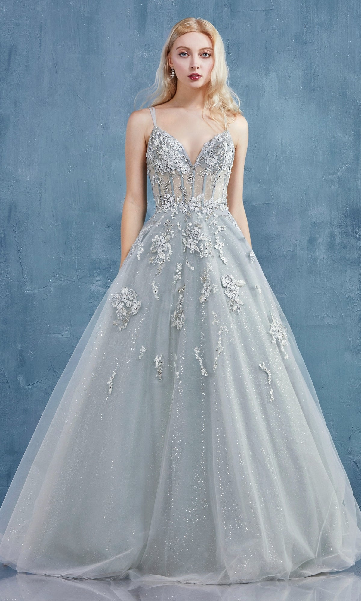 A woman in the Sage Green Long Glitter Prom Dress A0892, featuring floral appliqués and a sheer bodice, stands against a textured blue wall.